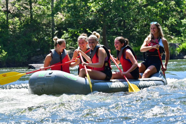 Five people wearing life jackets, paddling in an inflatable raft on a river.