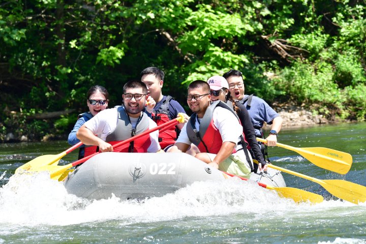 Group of people rafting on a river, smiling and holding paddles, surrounded by green trees.