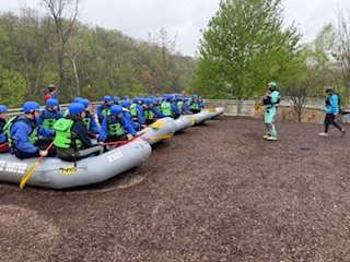 Group preparing for rafting, sitting in rafts on shore, wearing helmets and life vests.