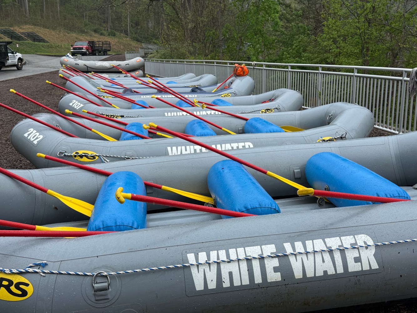 Gray inflatable rafts with blue seats and red paddles lined up on a gravel area.