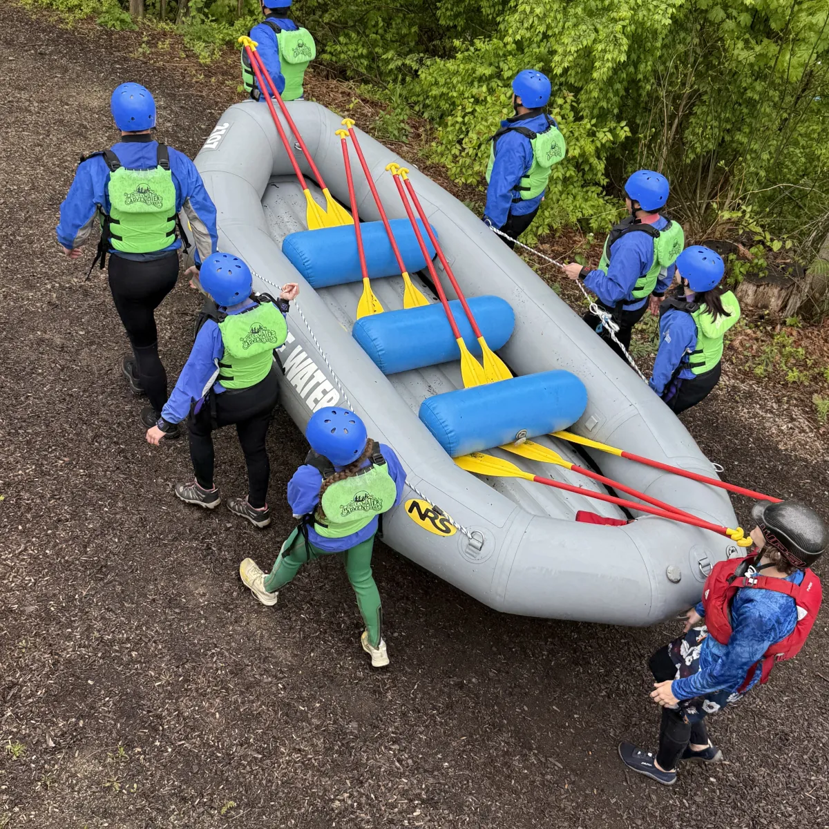Group carrying inflatable raft on a forest path, wearing helmets and life vests.