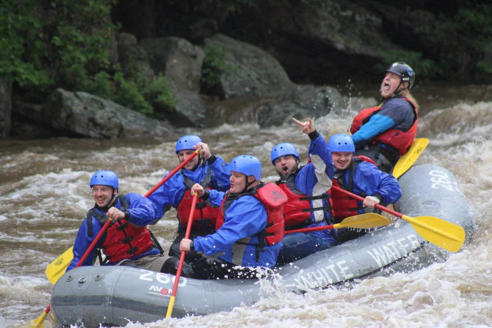 Group of six people in helmets rafting through rapids in an inflatable boat.