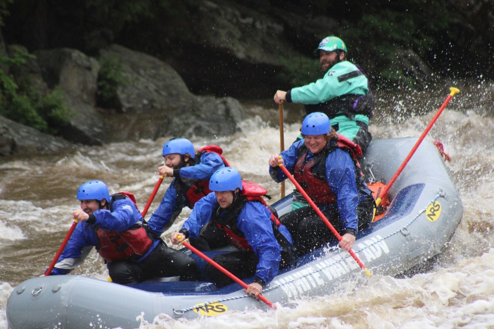Group white-water rafting in a river, wearing helmets and using paddles.