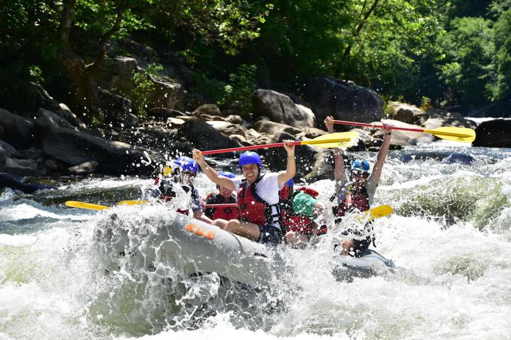 Group white-water rafting through rapids, paddles raised, wearing life jackets and helmets.