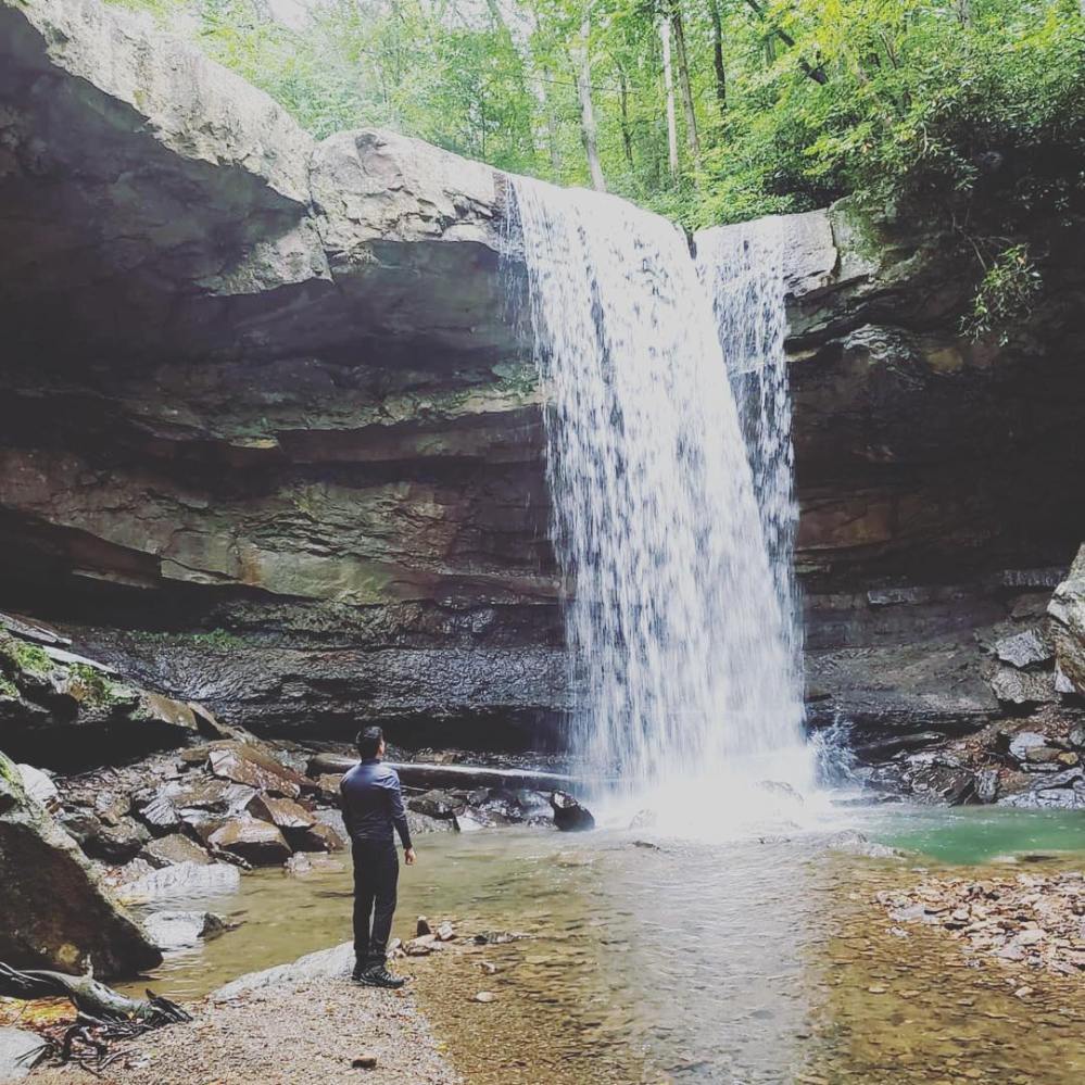 Person standing near a waterfall in a rocky forest setting.
