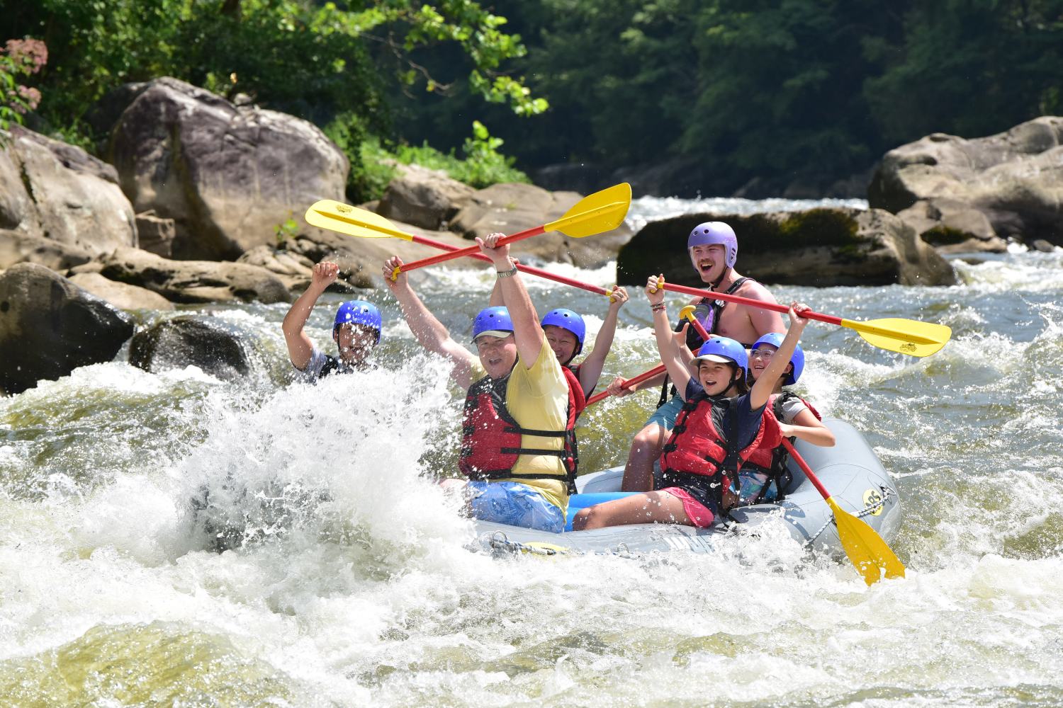 Group rafting on a river wearing helmets and life vests, paddling through rapids with raised paddles.